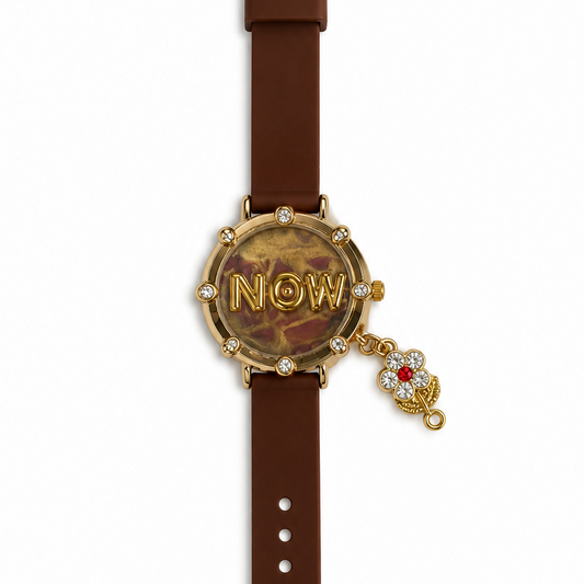 A women's wristwatch with a brown leather strap, a gold-tone round case, rhinestone accents, and a dangling charm featuring the word 'NOW' and a red jewel, shown on a plain white background.